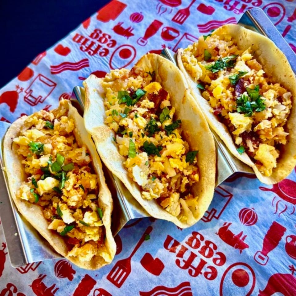 Three breakfast tacos with scrambled eggs, bacon, and cilantro, served on branded paper.