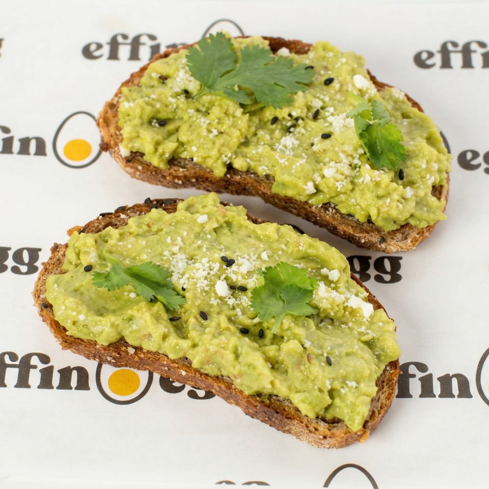 Two avocado toasts with cilantro, cheese, and black sesame seeds on Effin Egg branded paper.
