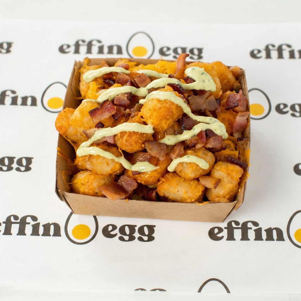 Loaded tater tots with cheese, bacon, and a creamy green sauce in a cardboard tray on an Effin Egg branded surface.